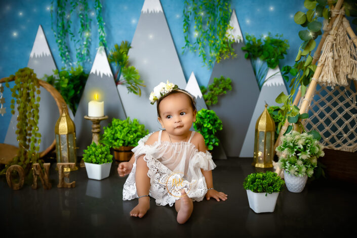 A baby girl in a white dress sitting on a floor with plants and a mountain backdrop captured by Delhi NCR's best maternal photographer Meghna Rathore.