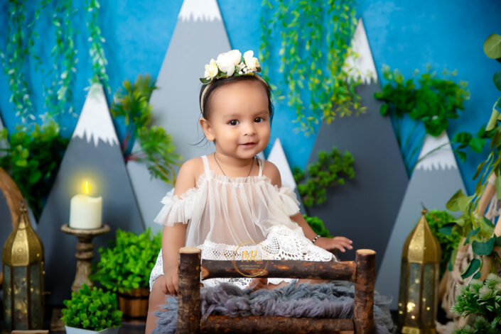 A baby girl in a white dress sitting on a wooden bed against a blue background containing mountain captured by Delhi NCR's best maternal photographer Meghna Rathore.