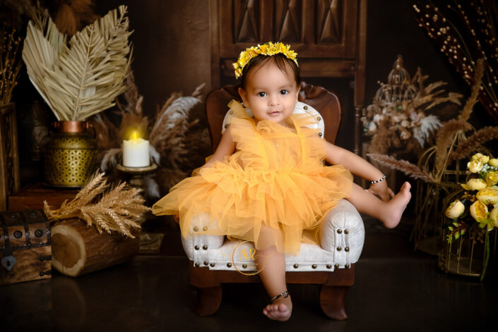 A baby girl in a yellow dress sitting on a wooden chair captured by Delhi NCR's best maternal photographer Meghna Rathore.