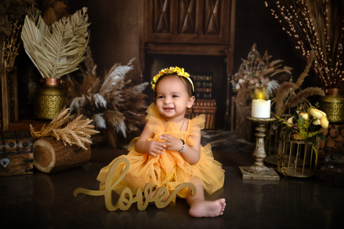 An infant girl in a yellow dress sitting on a wooden floor captured by Delhi NCR's best maternal photographer Meghna Rathore.
