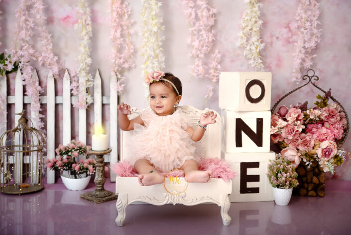 An adorable infant girl sitting on a white couch against pink backdrop with the number "one" displayed prominently captured by Delhi NCR's best maternal photographer Meghna Rathore.