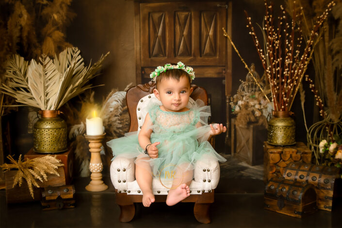 The child wears a flower crown and teal fabric, surrounded by ornate decor sitting on a white chair captured by Meghna Rathore Delhi NCR, Haryana best maternity and child photographer.