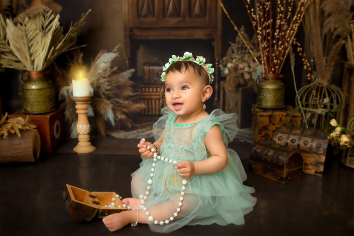 A child in a teal dress and floral headband holds pearls, surrounded by vintage, rustic decor and a lit candle, captured by Meghna Rathore Delhi NCR, Haryana best maternity and child photographer.