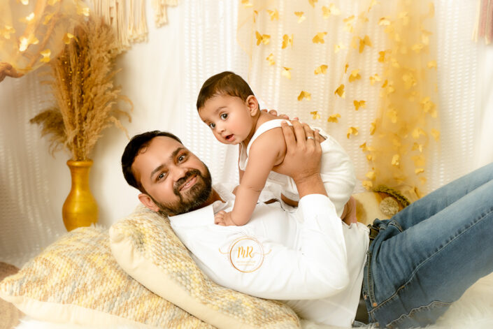 Father with his six month baby boy, both playing on cough surrounded with golden flask and golden curten having golden butterflies captured by Meghna Rathore, Delhi NCR's best maternity and child photographer.