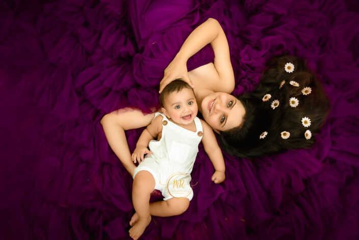 A smiling baby in white dress with her mother besides him in purple gown in a mom and me setup captured by Meghna Rathore, Delhi NCR's best maternity and child photographer.