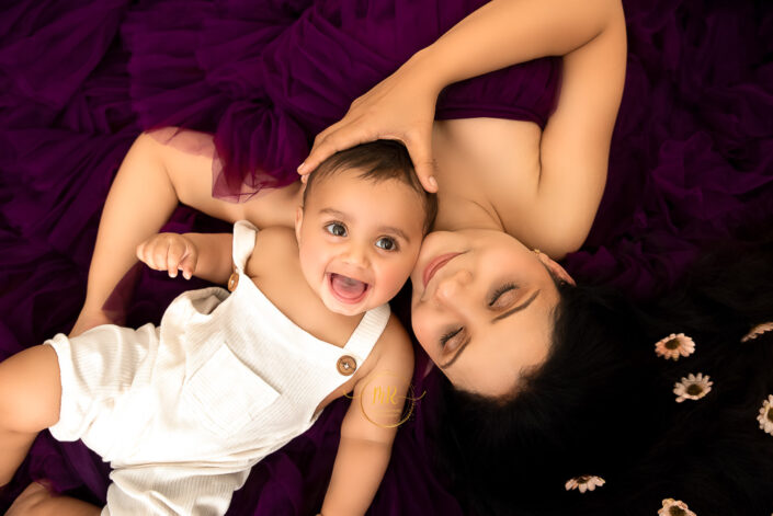 A baby in white dress with her mother besides him in purple gown in a mom and me setup captured by Meghna Rathore, Delhi NCR's best maternity and child photographer.
