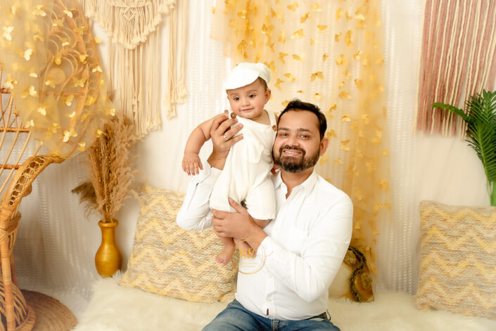 A child wearing white cap and white dress siting on his father shoulder in as elegant golden theme surrounding captured by Meghna Rathore, Delhi NCR's best maternity and child photographer.
