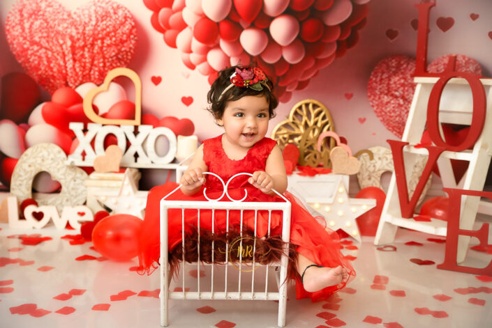 A baby girl in red dress and flowery tiara posing on aa small bed in a party themed background captured by Meghna Rathore best maternal and child photographer in Delhi, NCR.