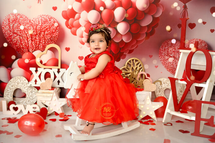 A cute little girl posing in her red dress riding wooden horse in a flowery background captured by Meghna Rathore best maternal and child photographer in Delhi, NCR.