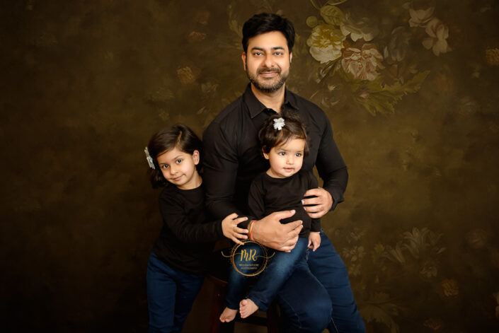 A father with his two daughters in black shirt and jeans in a dark brown flowery background captured by Meghna Rathore best maternal and child photographer in Delhi, NCR.