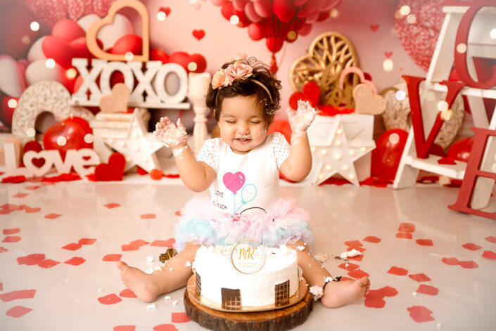 A baby in birthday dress eating and playing with white foster cake in a birthday background captured by Meghna Rathore best maternal and child photographer in Delhi, NCR.