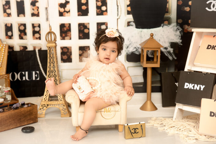 A cute little baby in tutu posing on a chair surrounded with french brand bags and miniature of Eiffel tower, captured by Meghna Rathore best maternal and child photographer in Delhi, NCR.