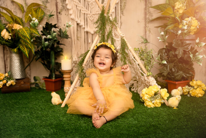 A smiling baby sitting on grass surrounded with chicks, green plants and yellow flowers, captured by Meghna Rathore best maternal and child photographer in Delhi, NCR.