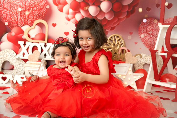 Two smiling and playing sisters in red dress in a party themed background posing for their photoshoot captured by Meghna Rathore best maternal and child photographer in Delhi, NCR.