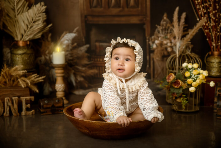 A baby in vintage attire sits in a wooden bowl, surrounded by antique decor, capturing the essence of innocence and nostalgia captured by by the best maternity and child photographer Meghna Rathore Delhi NCR, Haryana.