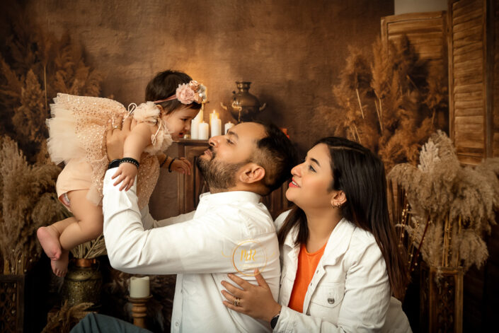 The image captures a tender moment between two parents and their baby in a vintage setting. The parents, faces are obscured for privacy, are interacting with the baby who is dressed in a light-colored outfit with ruffles and a flower headband. Captured by Delhi NCR best family and childhood photographer Meghna Rathore