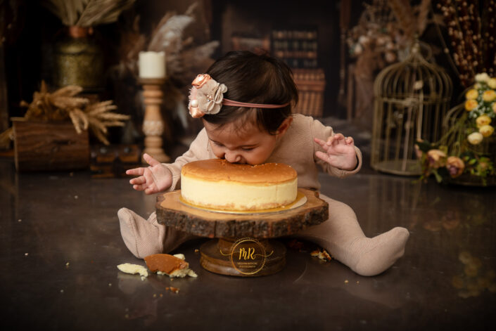 The image captures a joyful moment of a child participating in a cake smash. The child, dressed in a light-colored outfit, is seated amidst elegant decorations, reaching towards a cake on an ornate wooden slab by best kid photographer Meghna Rathore Delhi