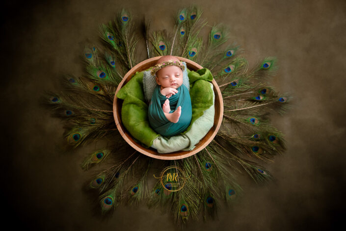 a one baby wrapped in green cloth, adorned with a floral crown, lies in a wooden bowl surrounded by peacock feather captured by Meghna Rathore Delhi NCR best child and maternity photographer