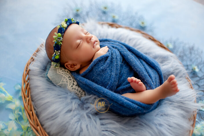 The image captures a serene and artistic scene of a baby wrapped in a blue cloth, lying in a basket surrounded by decorative elements, creating an aesthetic and peaceful atmosphere by meghna Delhi NCR best maternity and child photographer.
