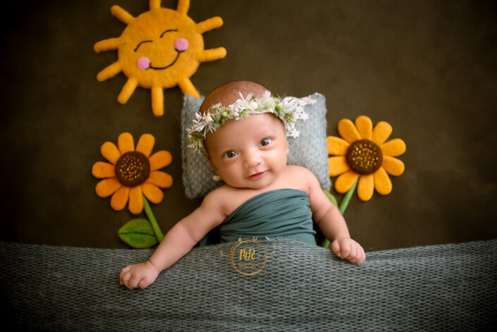 A baby with a flower crown lies on a textured surface, surrounded by crafted sunflowers and a smiling sun by Meghna Rathore the best child photographer in Delhi NCR, Haryana.