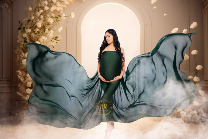 meghna rathore photography, delhi ncr, haryana, photoshoot by the best maternity photographer woman standing in front of a glowing doorway with vines of white flowers by the side and pillars on either of her, wearing a green satin dress with the backside positioned in the air at the angle of a butterfly