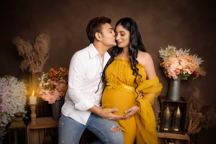 meghna rathore photography, delhi ncr, gururgram, haryana, maternity photoshoot by the best photographer, woman standing in front of a broown backdrop beside vases of flowers, lanterns and candles wearing a yellow maternity gown, husband kissing wife on her cheek
