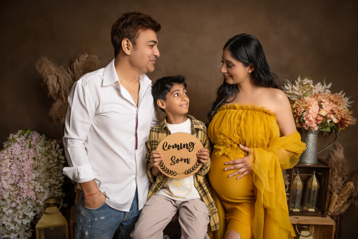 meghna rathore photography, delhi ncr, gurugram, haryana, maternity photoshoot by the best maternity photographer, family standing infront of a brown backdrop beside vases of flowers, shelves and lanterns, woman wearing a yellow maternity gown with her hand on her stomach, hsuband looking at his face wearing jeans and a white shirt, son sitting beside them holding a sign stating "coming soon"