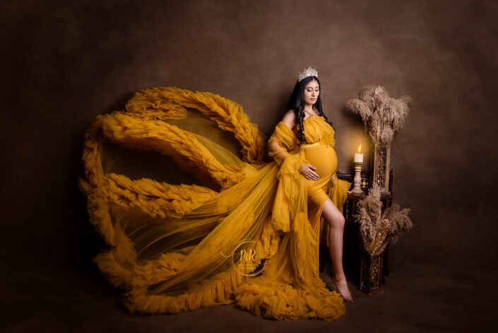 meghna rathore photography, delhi ncr, haryana, maternity photoshoot by the best maternity photographer woman standing infront of brown backdrop beside a bench with candles and vases of flowers, wearing a crown with jewels and a yellow maternity grown with the lower half positioned at an angle in the air like butterfly wings