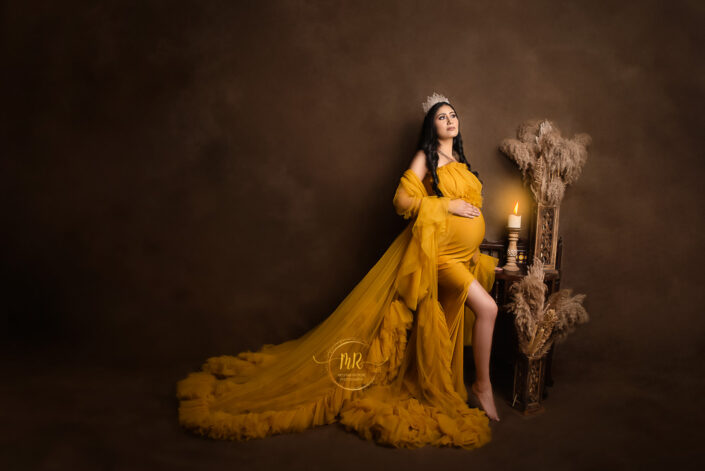 meghna rathore photography, delhi ncr, haryana, maternity photoshoot by the best maternity photographer, woman wearing a yellow dress with a trailling back and her hand on her stomach, standing in front of a painted brown backdrop beside to a bench with candles and vases of flowers