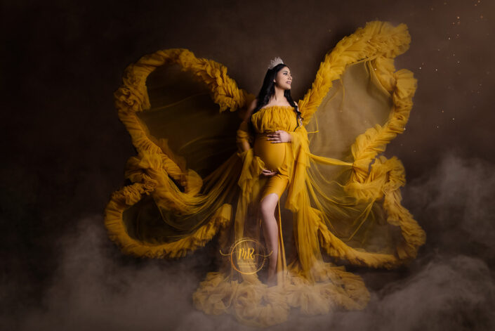 meghna rathore photography, Delhi ncr, Haryana, maternity photoshoot by the best maternity photographer, women standing infront of a painted brown backdrop with shimmers on the side and smoke encircling her legs and gown, woman wearing a yellow dress and a crown with jewels with her hand on her stomach, womans dress positioned at an angle like butterfly wings in the air