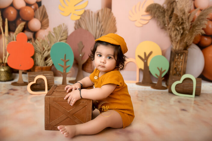A baby in a yellow outfit sits amidst whimsical forest-themed decorations, holding a wooden box, creating a playful, enchanting atmosphere of childhood wonder by the best maternity and Pre-Birthday child photographer Meghna Rathore Delhi NCR, Haryana.
