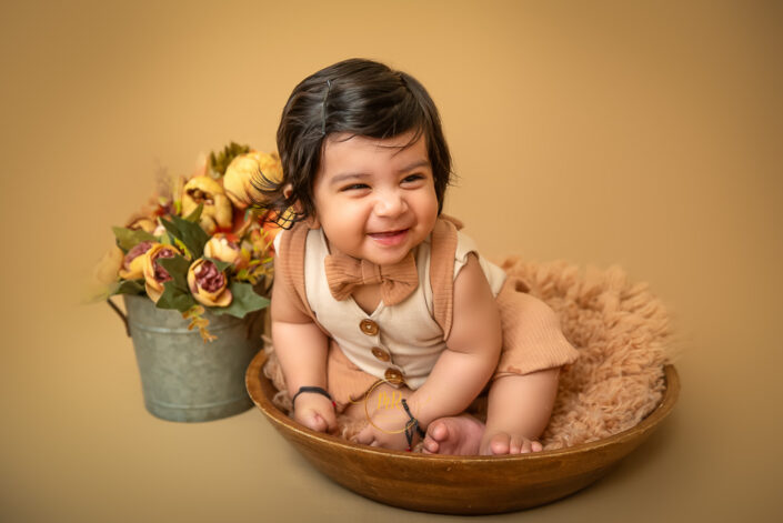 A cute baby boy in wooden bowl smiling wearing a bow surrounded by roses captured by Meghna Rathore Delhi NCR best maternity and child photographer.