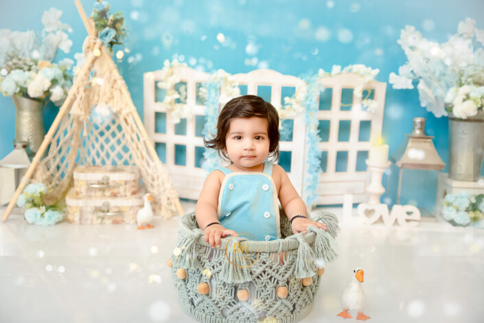 A child sits in a basket amidst a whimsical setting with flowers, a teepee, windows, and a ‘love’ sign, creating an enchanting atmosphere by the best maternity and Pre-Birthday child photographer Meghna Rathore Delhi NCR, Haryana.