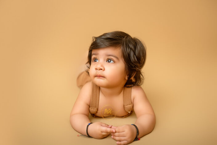 A child wearing suspenders and bracelets, sits against a monochromatic background. The image captures a child in suspenders, evoking a sense of innocence and nostalgia by the best maternity and Pre-Birthday child photographer Meghna Rathore Delhi NCR, Haryana.
